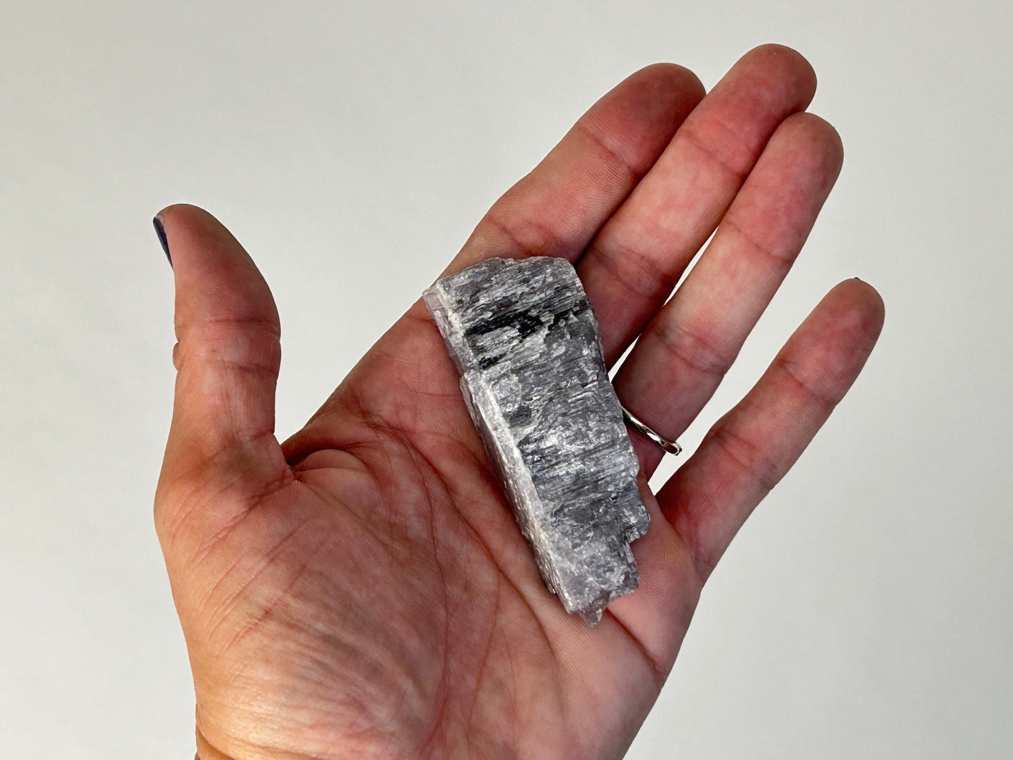 A hand holding a rough, metallic gray Energized Natural Purple Anhydrite specimen against a plain light background.