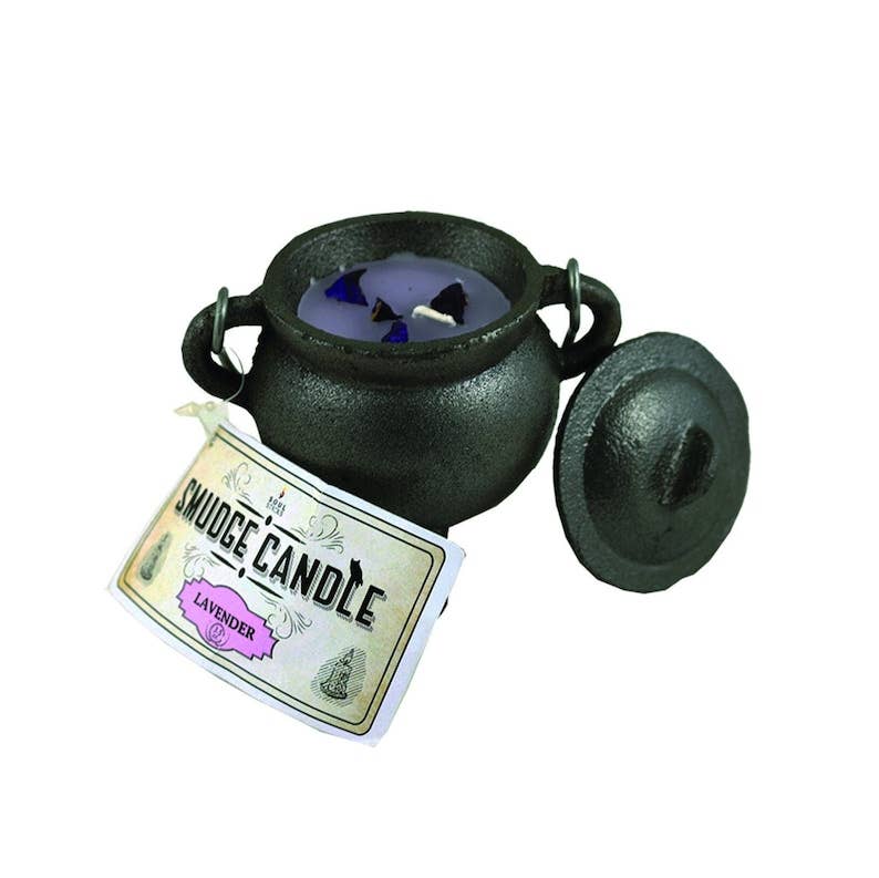 A cast-iron cauldron-shaped candle with its lid off, showing lavender-colored wax and blue glass chips inside. A paper tag attached to the front reads 'SMUDGE CANDLE' and 'LAVENDER'.