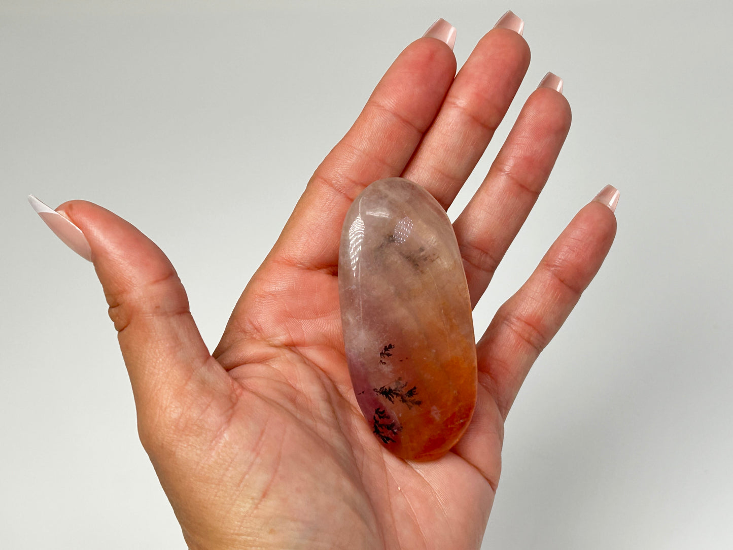 Hand holding a Polished, oval-shaped light-brown Dendritic Fluorite mineral palm stone with brown and orange colored markings reminiscent of a landscape healing crystal gemstone for meditation and energy work with white background.