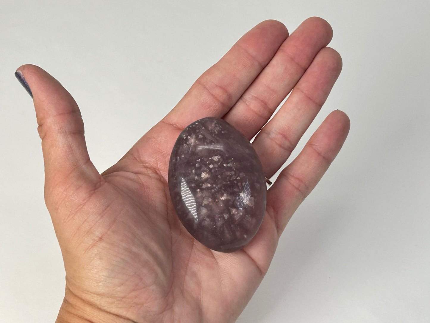 A hand holding a polished oval-shaped Purple Fluorite with Mica Palm Stone with a dark gray and brown speckled pattern, shown against a plain light gray background.