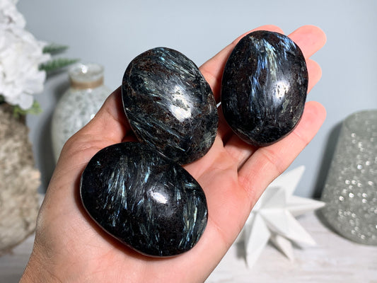 Three polished Anthophyllite in Eudialyte with Pyrite Palm Stone Crystal  held in a person's hand, showing iridescent blue and white flashes, with a softly blurred decorative background.