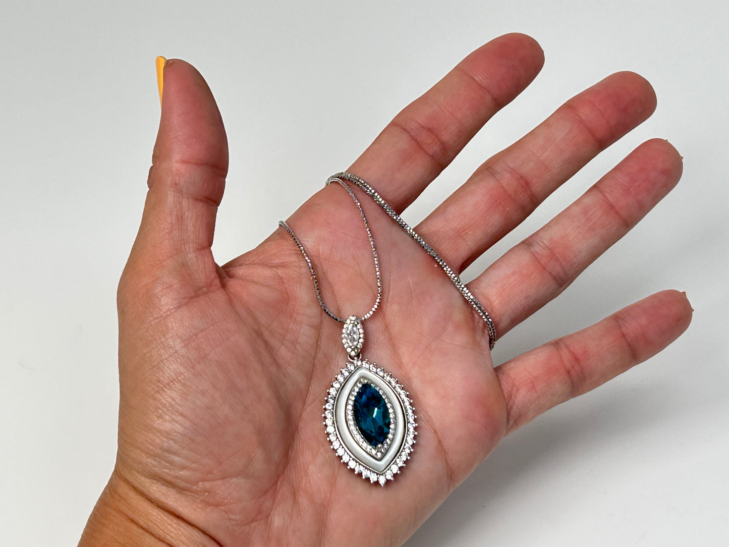 Hand holding a silver necklace with a Blue Topaz Eye Pendant with Cubic Zirconia in Sterling Silver for meditation and energy work with white gray floral background