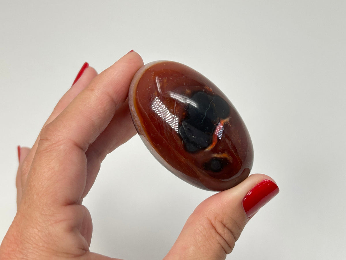 A hand with red nail polish holds a polished oval Carnelian Crystal Shiva featuring a rich reddish-brown body with prominent black and white banding.