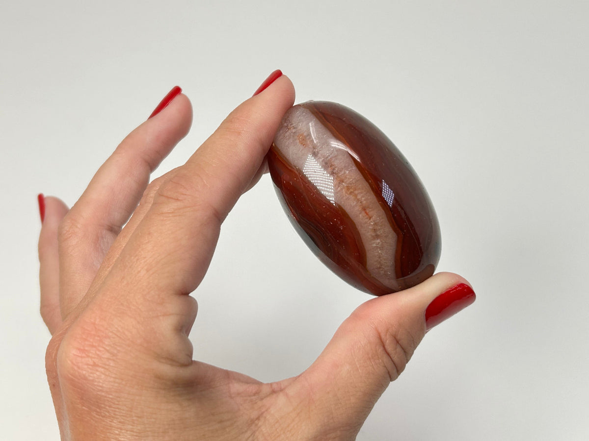 A hand with red nail polish holds a polished oval Carnelian Crystal Shiva against a plain light gray background.