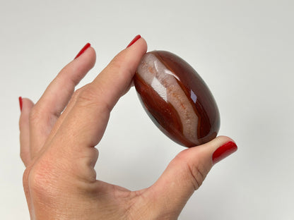 A hand with red nail polish holds a polished oval Carnelian Crystal Shiva against a plain light gray background.