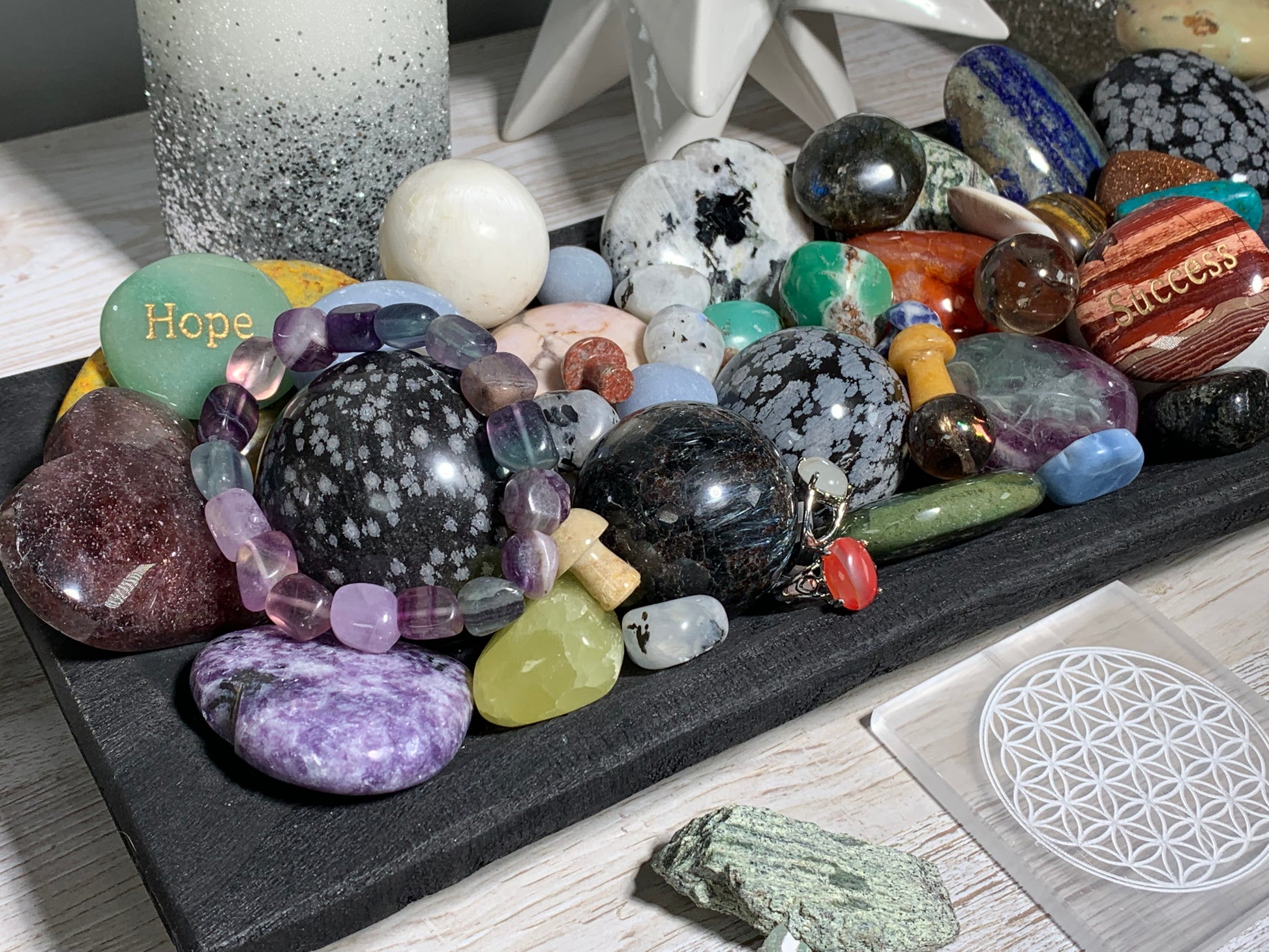 A collection of polished and raw gemstones, crystals, beads and rings arranged on a black slate tray, featuring various colors and patterns including moonstone, agate, quartz, and a green stone engraved with 'Hope'.