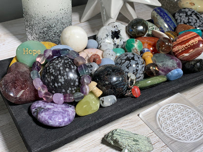 A collection of polished and raw gemstones, crystals, beads and rings arranged on a black slate tray, featuring various colors and patterns including moonstone, agate, quartz, and a green stone engraved with 'Hope'.