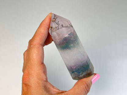 Hand holding a Dendritic Fluorite Tower gemstone on table top with white background