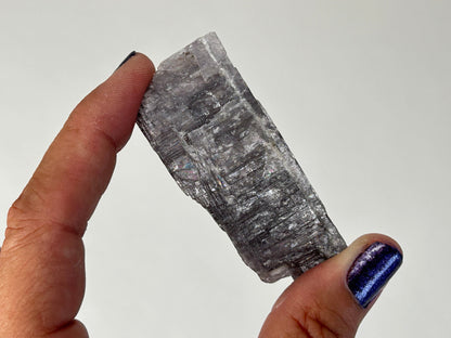 A hand holding a raw, rough translucent gray Energized Natural Purple Anhydrite against a plain light gray background.