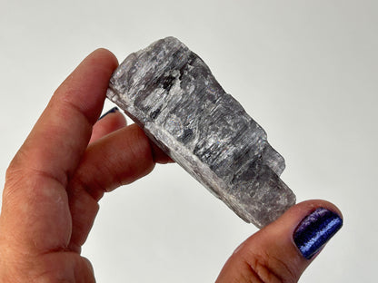 A hand holding a raw, rough gray Energized Natural Purple Anhydrite specimen against a plain light gray background.