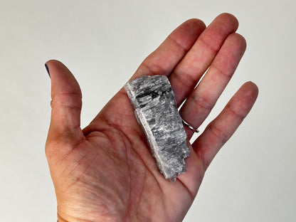 A hand holding a rough, metallic gray Energized Natural Purple Anhydrite specimen against a plain light background.
