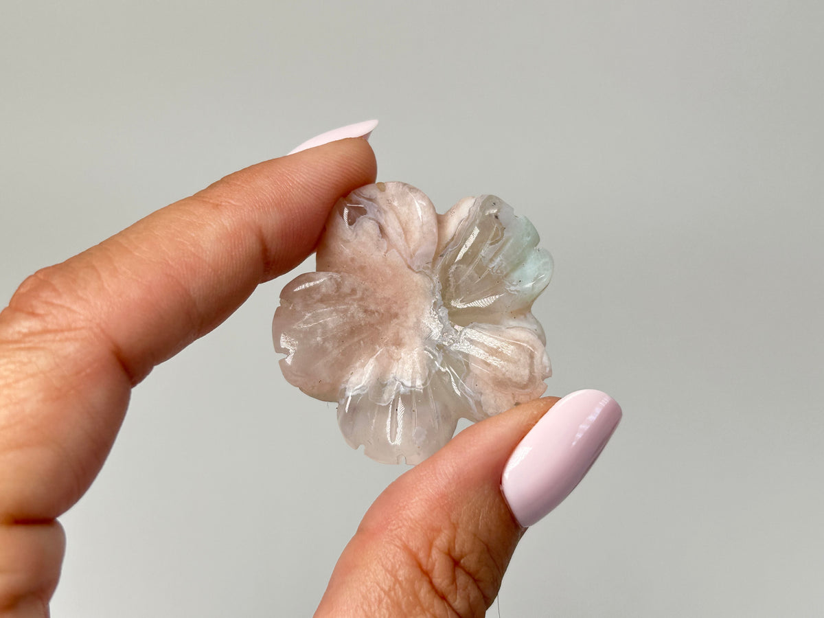 A hand with light pink manicured nails holds a small, translucent flower-shaped gemstone or crystal pendant against a plain light gray background.