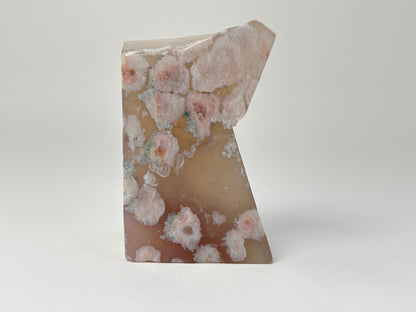 A polished natural agate stone sculpture with a distinctive abstract, stepped shape, featuring a marbled pattern of pink, white, and brown mineral deposits on a light gray background.