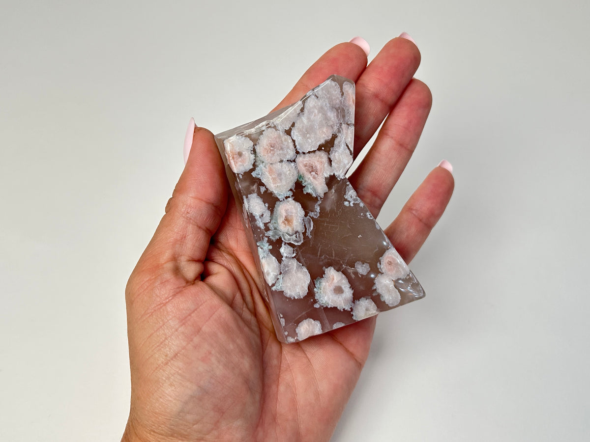 A hand holding a polished rectangular stone with a translucent taupe-brown body and numerous white to pale pink floral inclusions, shown against a plain light gray background.