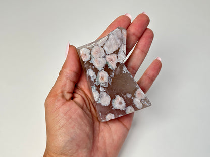 A hand holding a polished rectangular stone with a translucent taupe-brown body and numerous white to pale pink floral inclusions, shown against a plain light gray background.