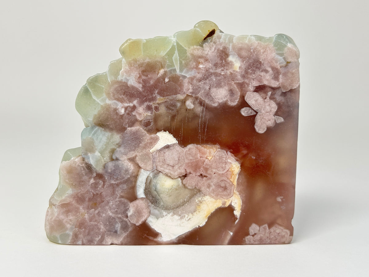 A carved natural agate stone plaque featuring pinkish-brown floral clusters, pale green edges, and a white crystalline center, photographed on a neutral light gray background.