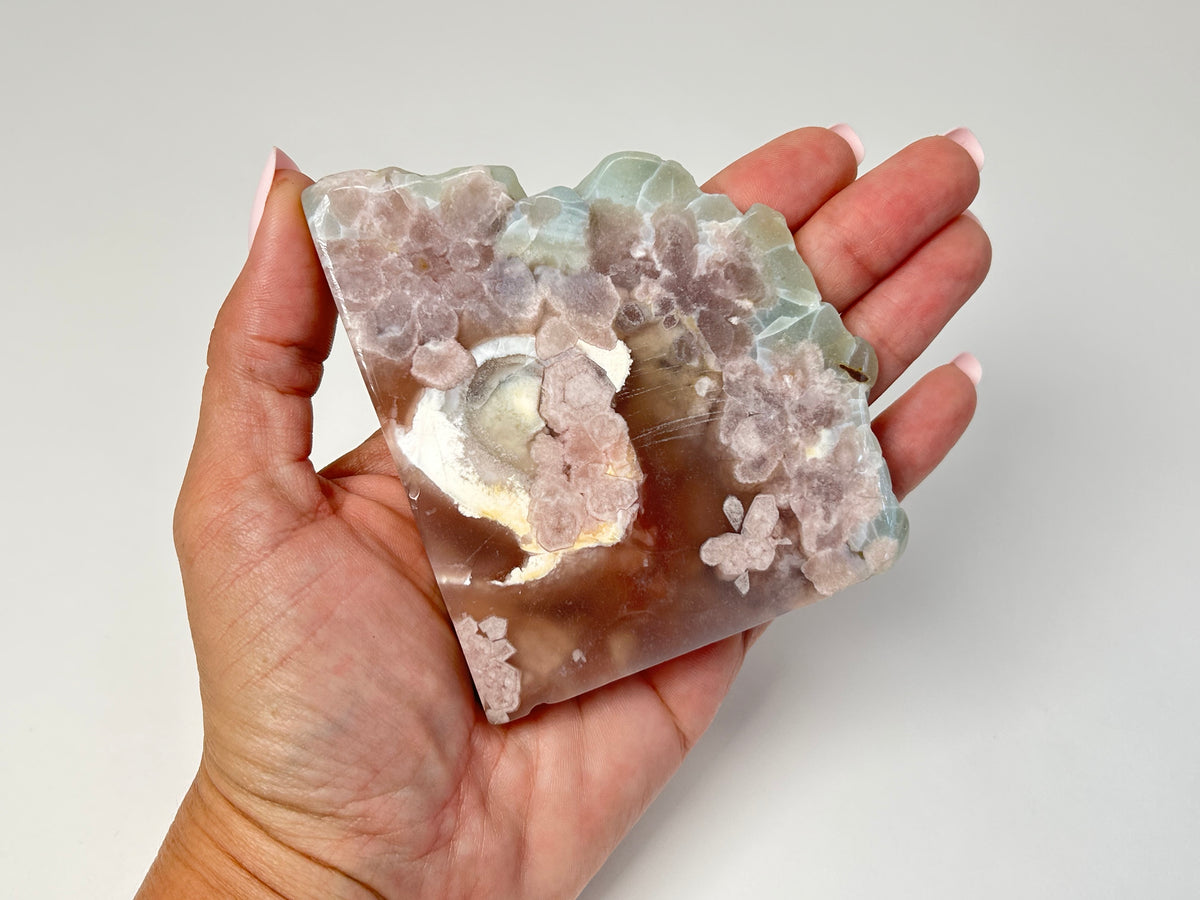 A hand holding a polished agate crystal specimen with pink, white, and light green formations against a plain light gray background.