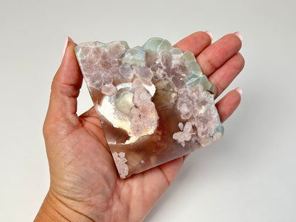 A hand holding a polished agate crystal specimen with pink, white, and light green formations against a plain light gray background.