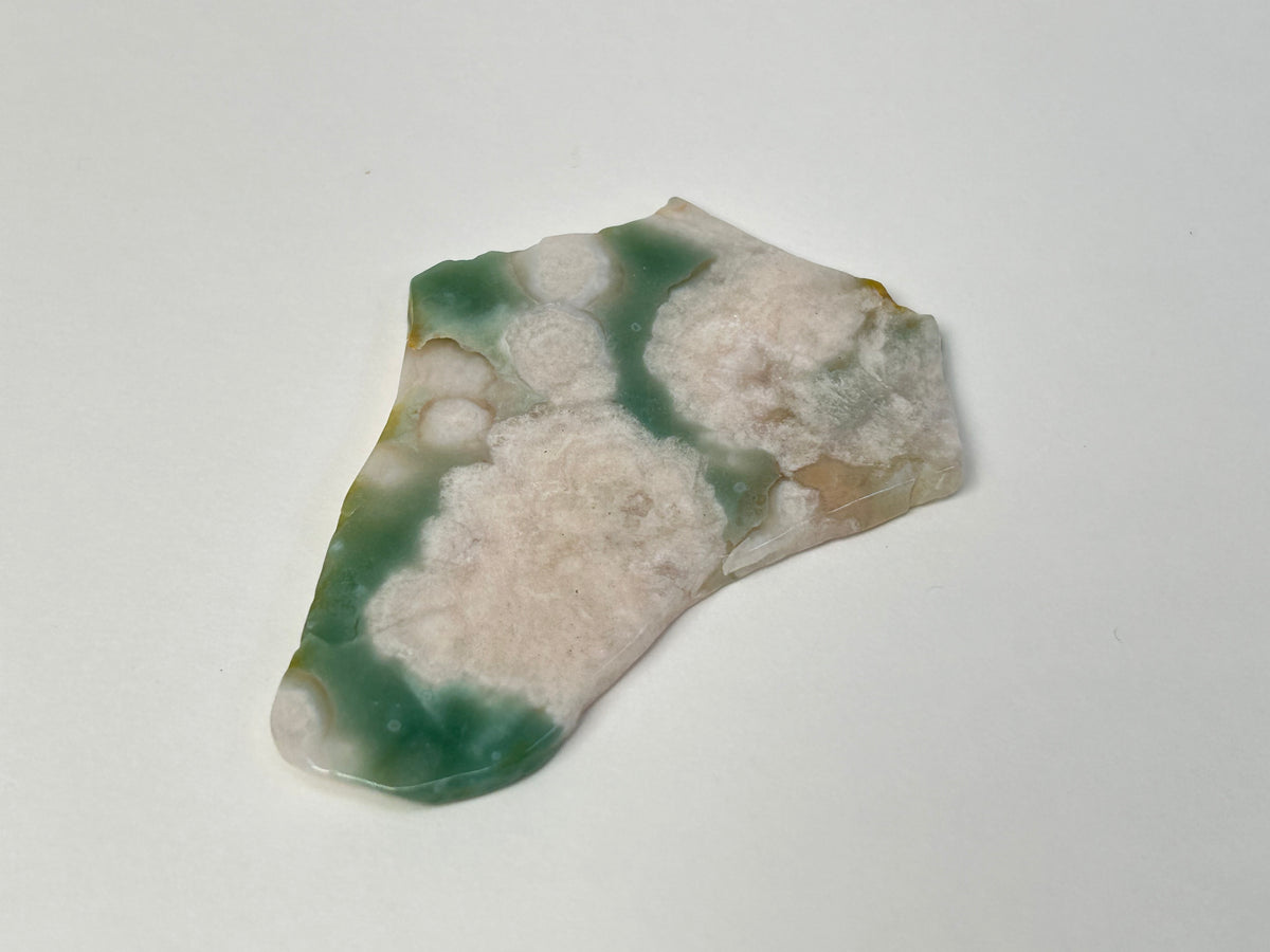 A polished natural stone slice with a marbled pattern of green, cream, and beige, photographed on a plain light gray background.
