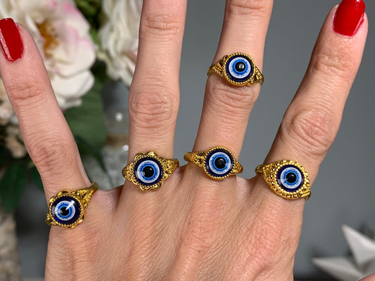 A close-up of a hand wearing five gold rings, each featuring a blue and white evil eye design with a black center, set against a soft blurred background with flowers.