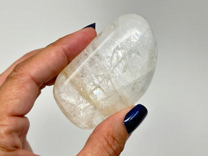 A hand with dark blue nail polish holds a High-Vibrational Natrolite Freeform against a plain light gray background.