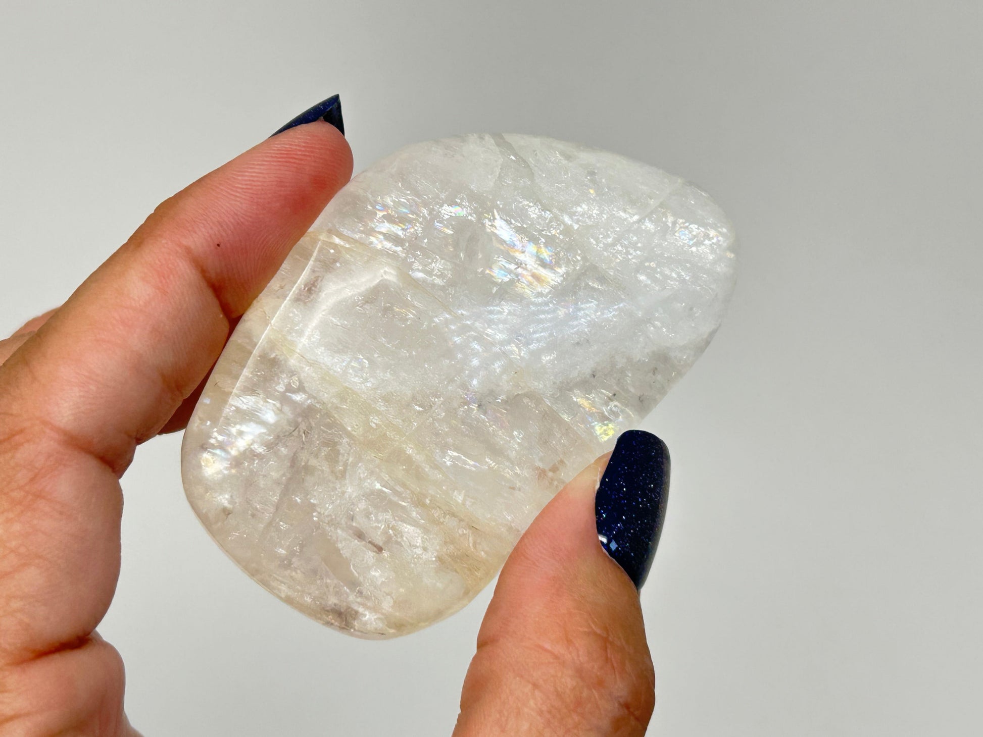 A hand with dark glittery nail polish holds a polished, irregular-shaped white High-Vibrational Natrolite Freeform against a plain light gray background.
