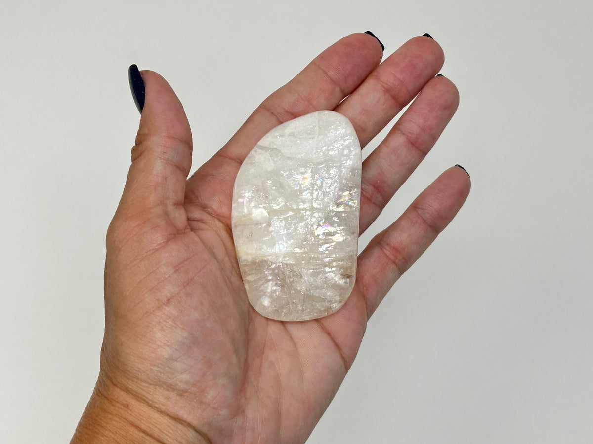 A hand with dark nail polish holds a polished, High-Vibrational Natrolite Freeform against a plain light gray background.