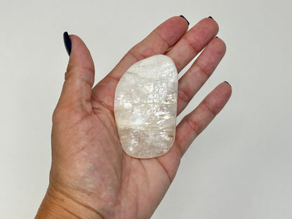 A hand with dark nail polish holds a polished, High-Vibrational Natrolite Freeform against a plain light gray background.