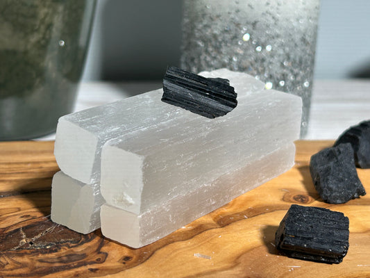 Four white Selenite sticks arranged in two pairs on a wooden surface with a piece of Black Tourmaline crystal on top of the sticks.
