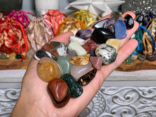 A hand holding a collection of polished tumbled gemstones in various colors, with colorful drawstring pouches and decorative items blurred in the background.