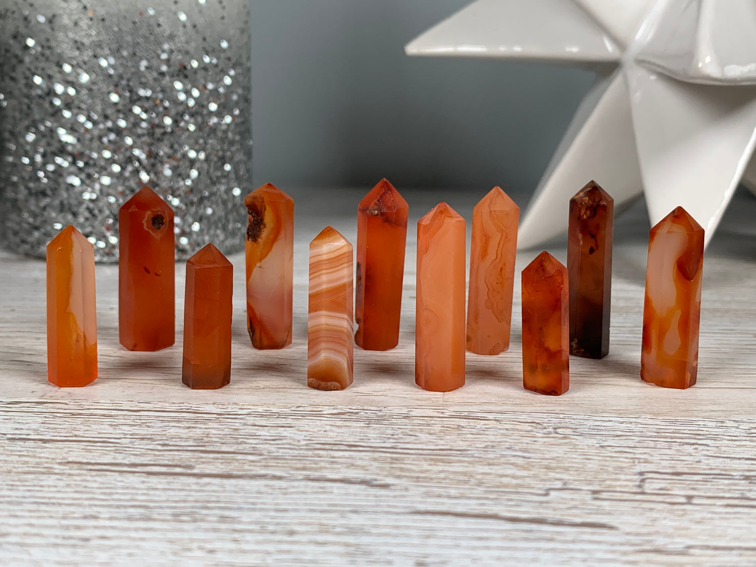 A set of ten polished natural Mini Carnelian Crystal Towers in various orange, red, and brown tones, arranged on a light gray wooden surface with decorative background props.