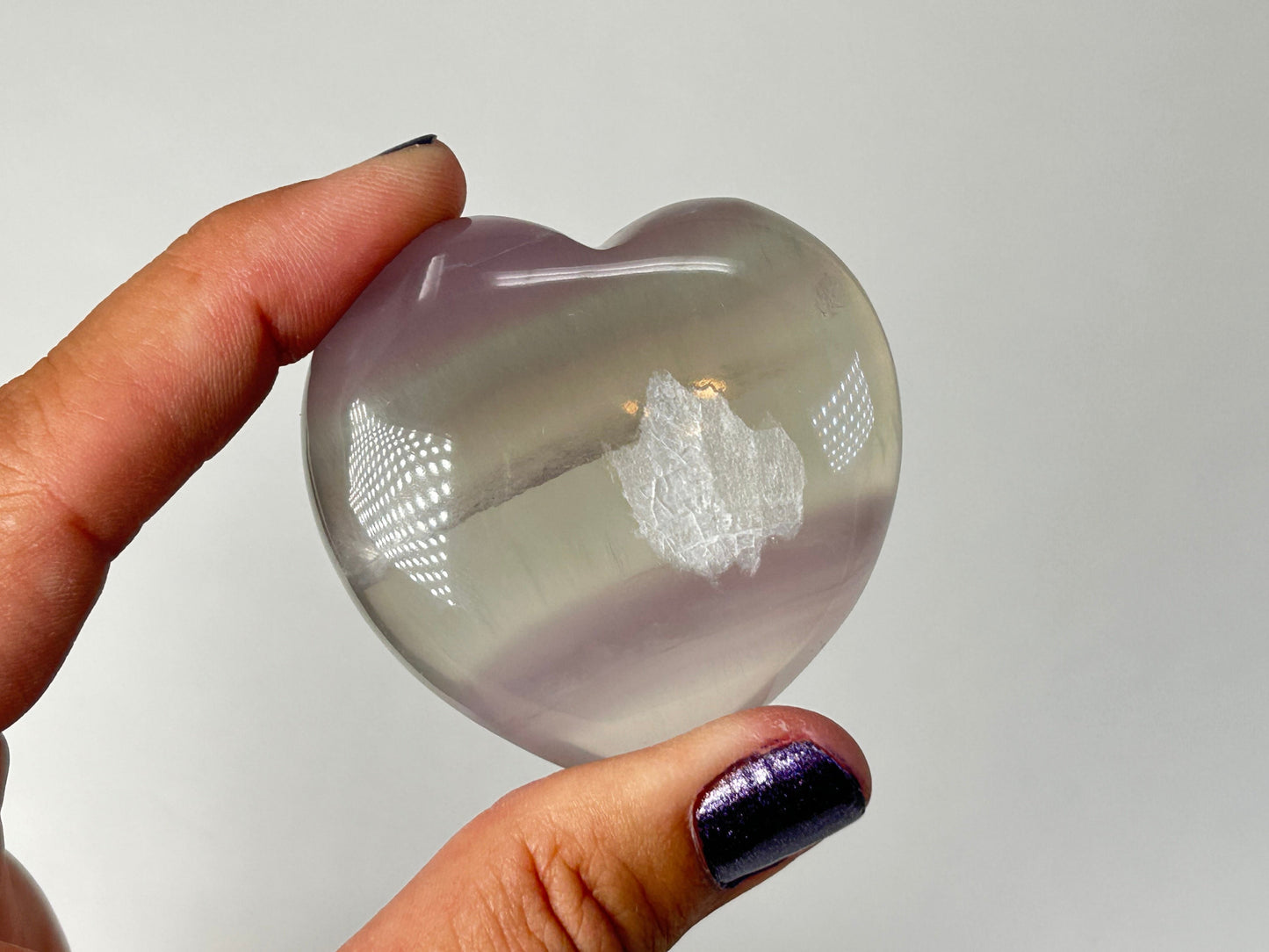 A hand holding a polished, Naturally Striped Fluorite Heart  with subtle lavender-purple tones and a white inclusions pattern in the center, set against a plain light gray background.