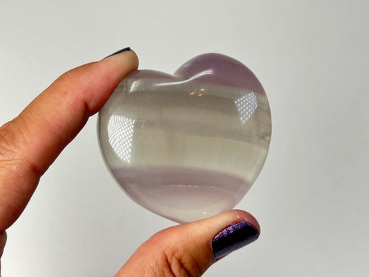 A hand holding a polished, Naturally Striped Fluorite Heart with subtle lavender-purple banding near the top, set against a plain light gray background.