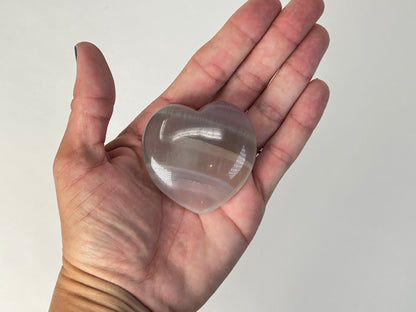 A translucent heart-shaped Naturally Striped Fluorite Heart held in a person's open palm against a plain light background.