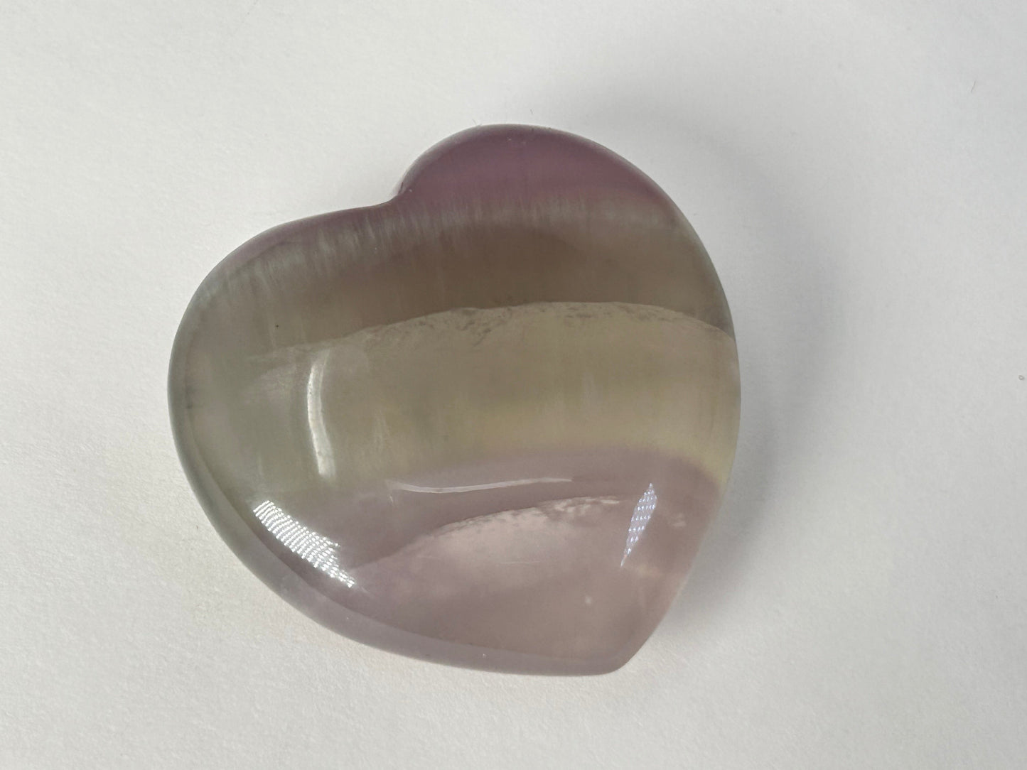 A heart-shaped polished Naturally Striped Fluorite Heart with a smooth, rounded surface, featuring a color gradient from purple at the top to a translucent grayish-green in the middle and a soft lavender-pink at the bottom, resting on a plain light gray surface.