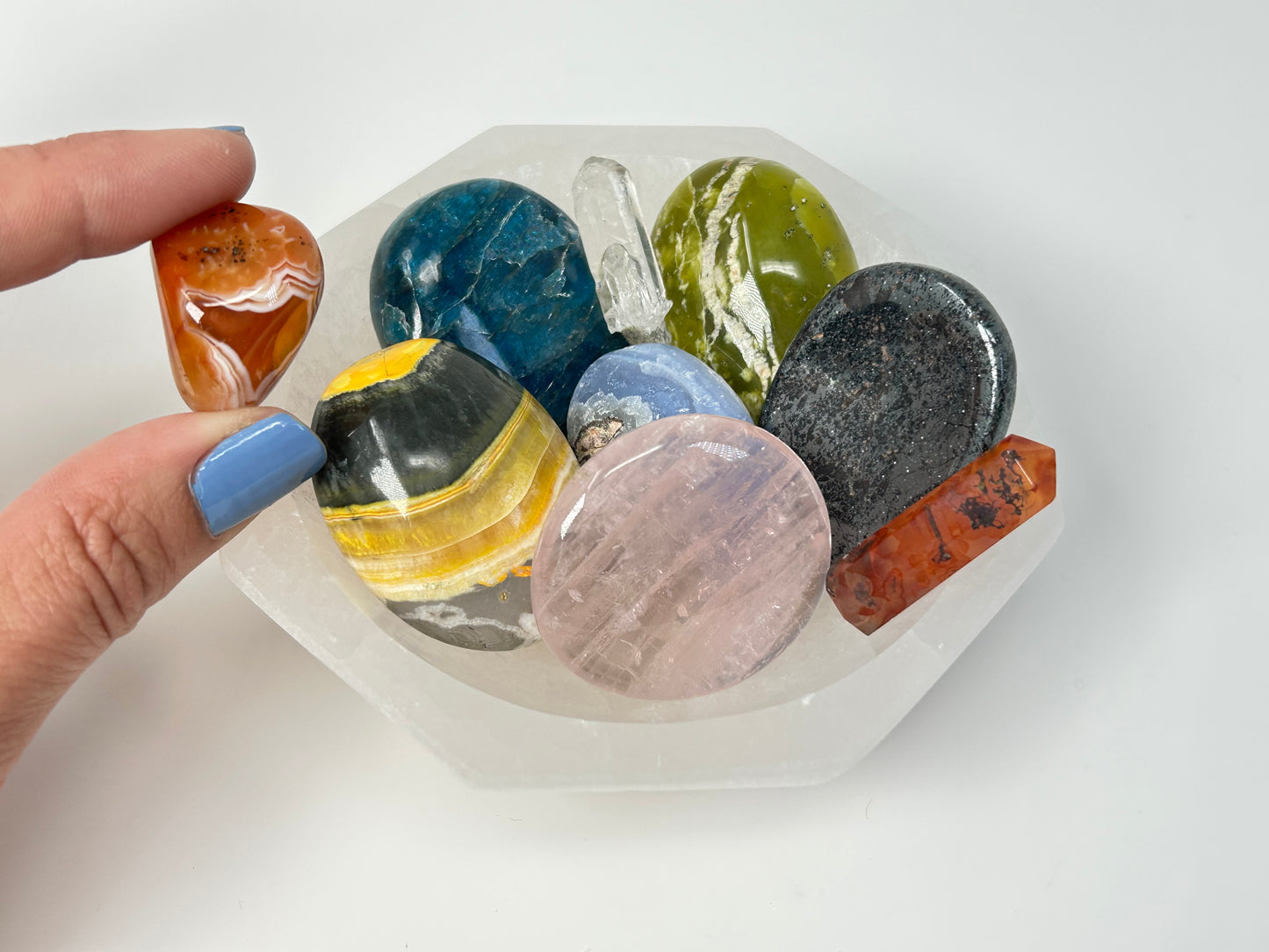 Hand holding a collection of colorful stones in an Octagon Selenite Bowl from Morocco for energy healing on table with white background.