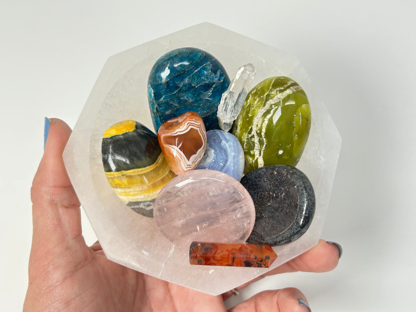 Hand holding a Octagon Selenite Bowl from Morocco for meditation and energy work with various colorful stones inside on a white background