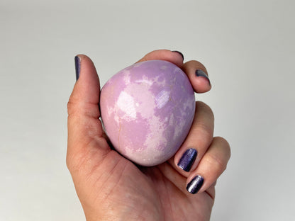 A hand with dark purple glitter nail polish holds a pastel lavender and white marbled Phosphosiderite Compassion Pebble against a plain light gray background.