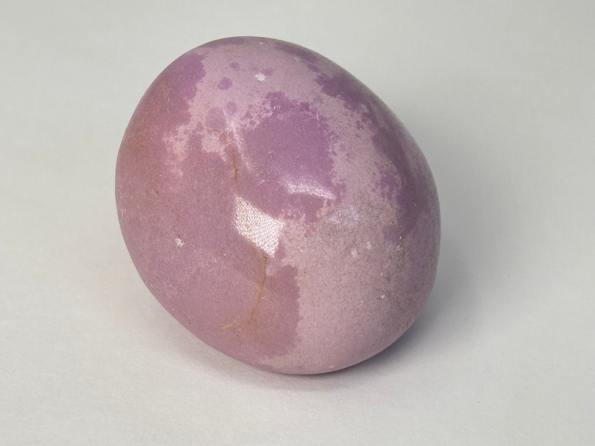 A smooth, polished oval Phosphosiderite Compassion Pebble in a dusty lavender-purple color with natural mottled patches and a small vertical fissure, photographed on a neutral light gray background.