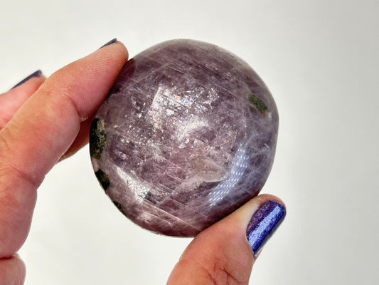 A hand with purple glitter nail polish holds a polished oval Purple Anhydrite Palm Stone showing lavender and gray tones with white striations, against a plain white background.