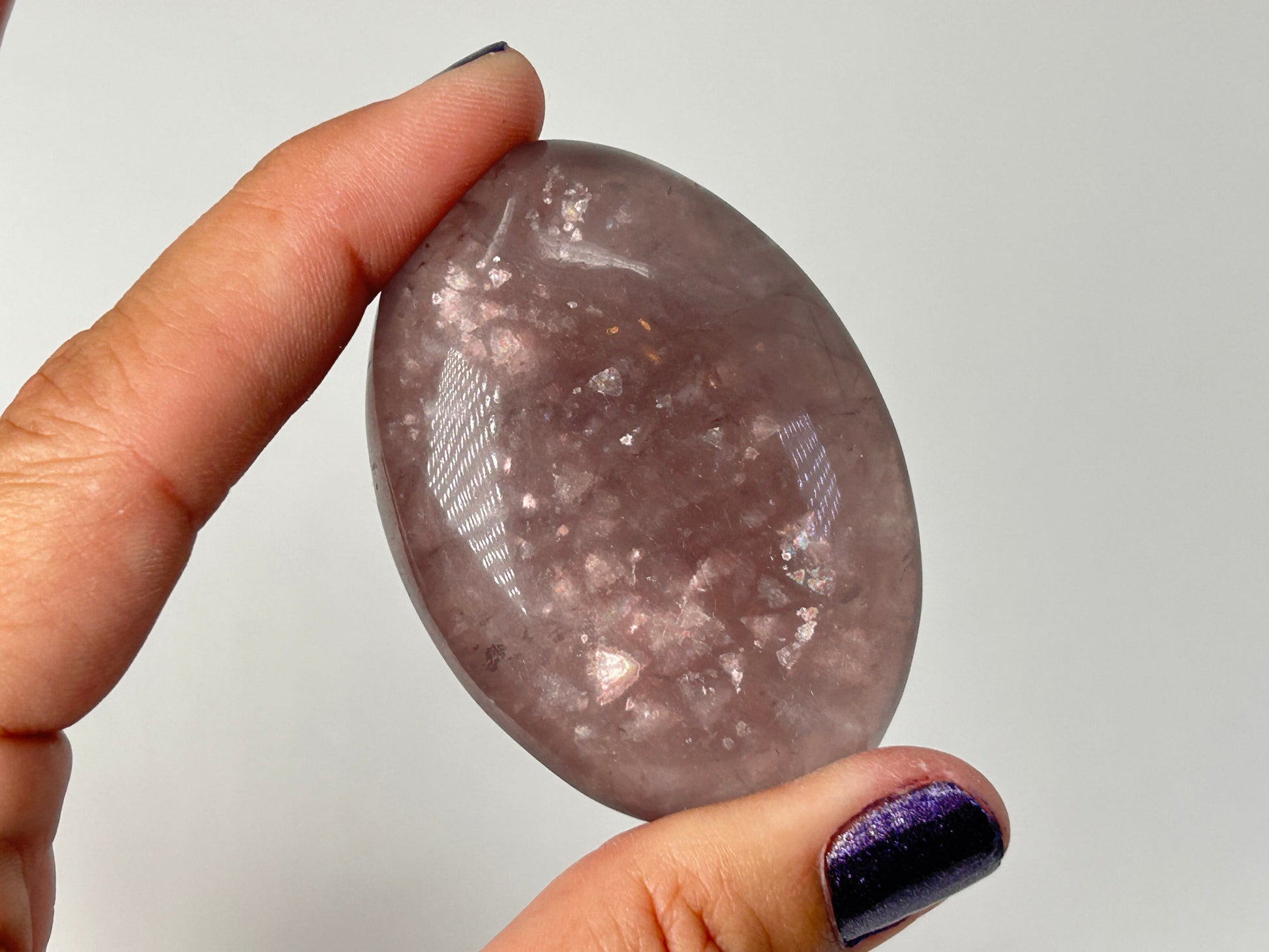 A hand holding a polished oval Purple Fluorite with Mica Palm Stone against a plain light gray background.