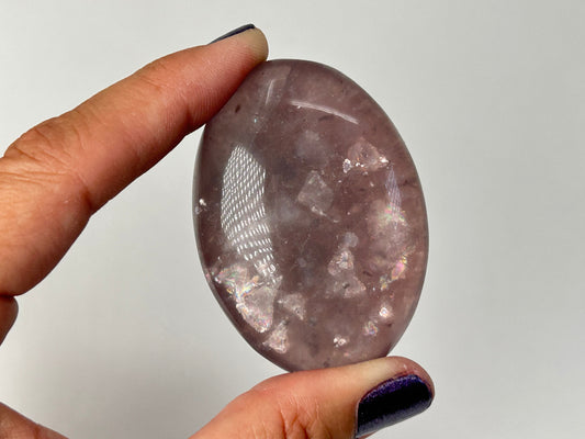 A hand holding a polished oval-shaped pinkish-purple Purple Fluorite with Mica Palm Stone, against a plain light gray background.