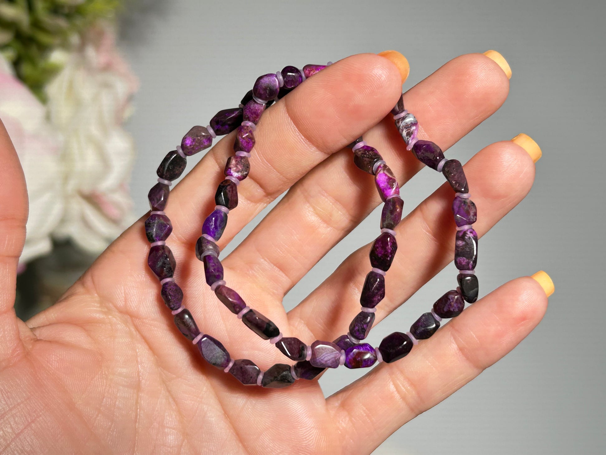 Hand holding two Purple Sugilite Bracelets with floral background.