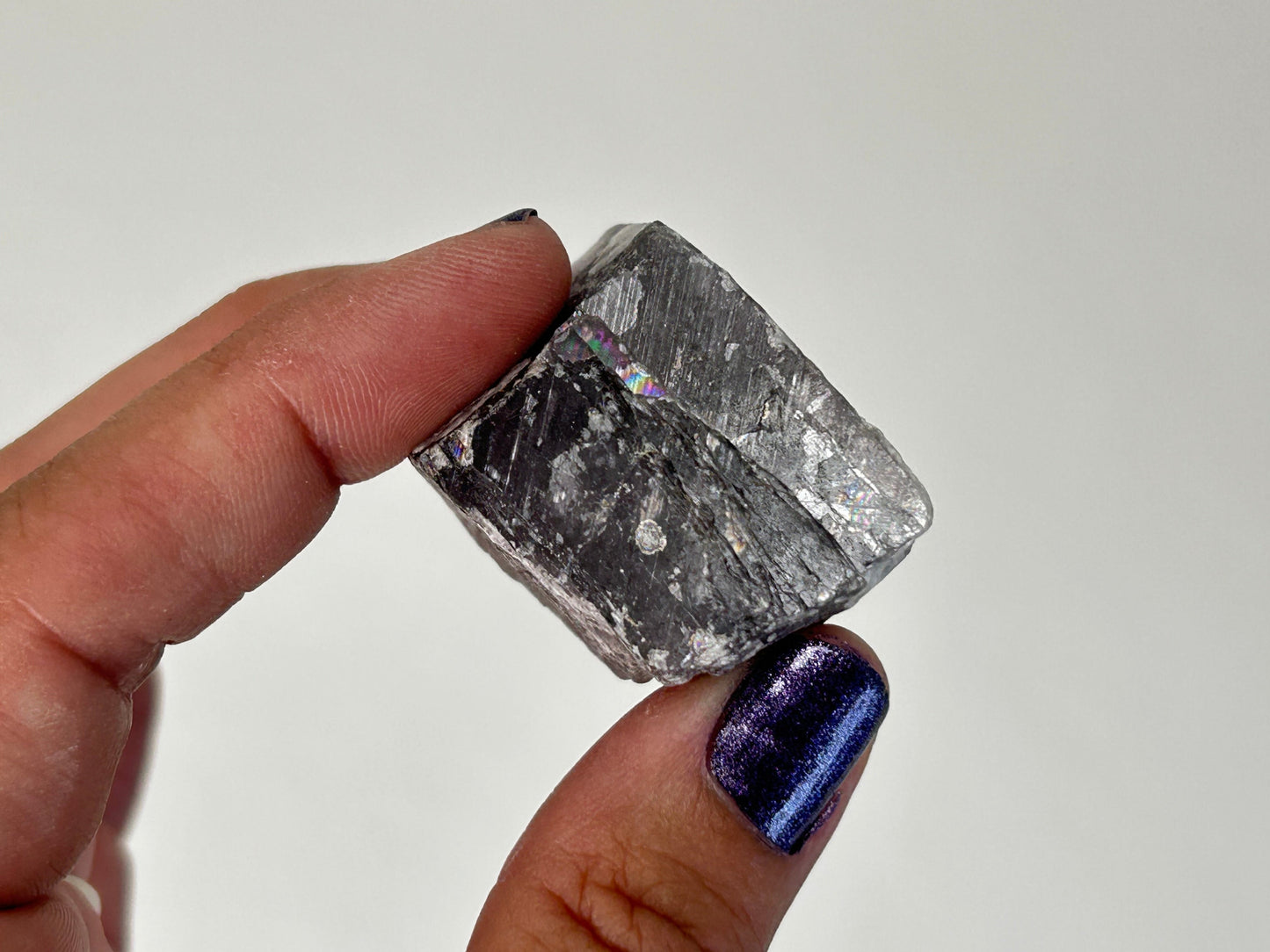 A hand holding a rough, metallic gray rectangular Radiant Natural Purple Anhydrite Crystal against a plain light background.