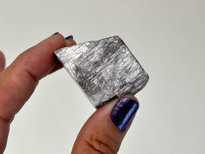 A hand with dark purple glitter nail polish holds a rough, metallic silver Radiant Natural Purple Anhydrite crystal against a plain light gray background