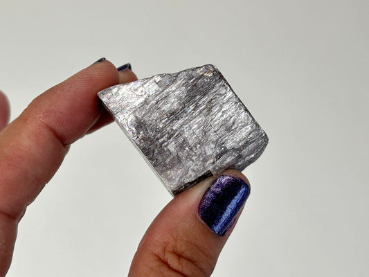A hand with dark purple glitter nail polish holds a rough, metallic silver Radiant Natural Purple Anhydrite crystal against a plain light gray background
