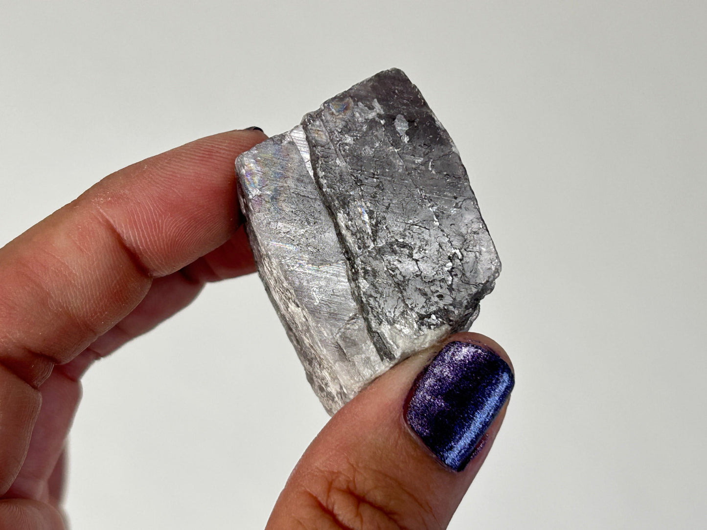 A hand holding a rough, metallic silver-gray Radiant Natural Purple Anhydrite Crystal specimen against a plain light background.