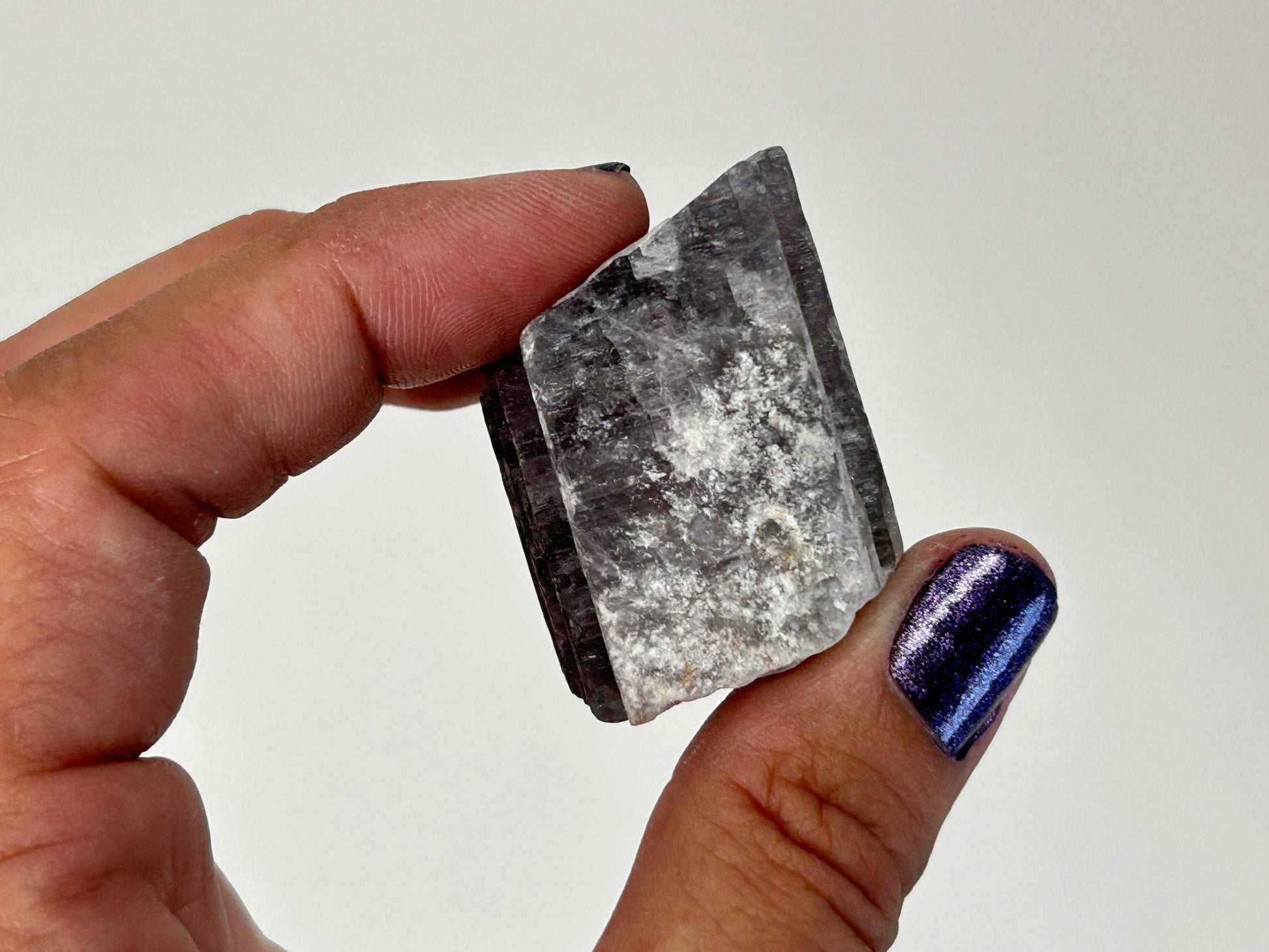A hand holding a Radiant Natural Purple Anhydrite Crystal with a jagged, unpolished edge. The crystal is primarily gray with white cloudy inclusions and dark charcoal sides.