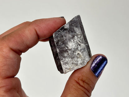 A hand holding a Radiant Natural Purple Anhydrite Crystal with a jagged, unpolished edge. The crystal is primarily gray with white cloudy inclusions and dark charcoal sides.