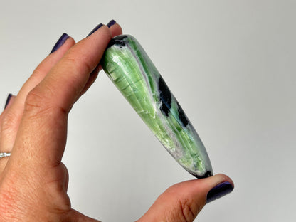 A hand with dark purple nail polish holds a polished Rare Green and Purple Kammererite Wand against a plain light gray background.